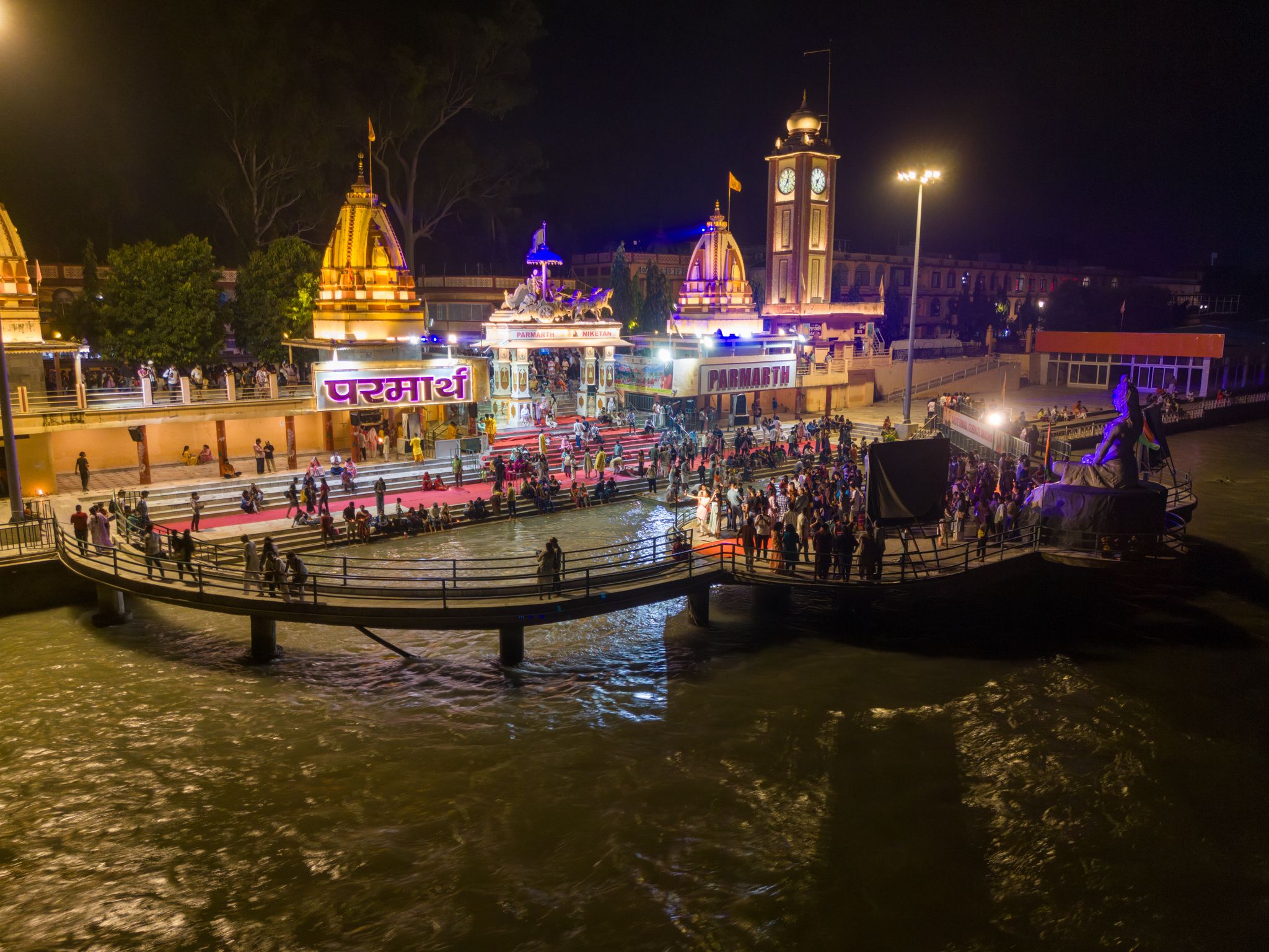 Aerial view at night of the lit ghat at Parmarth Niketan on the Ganga in Rishikesh, with temple domes, a clock tower and crowds gathered for the evening aarti.