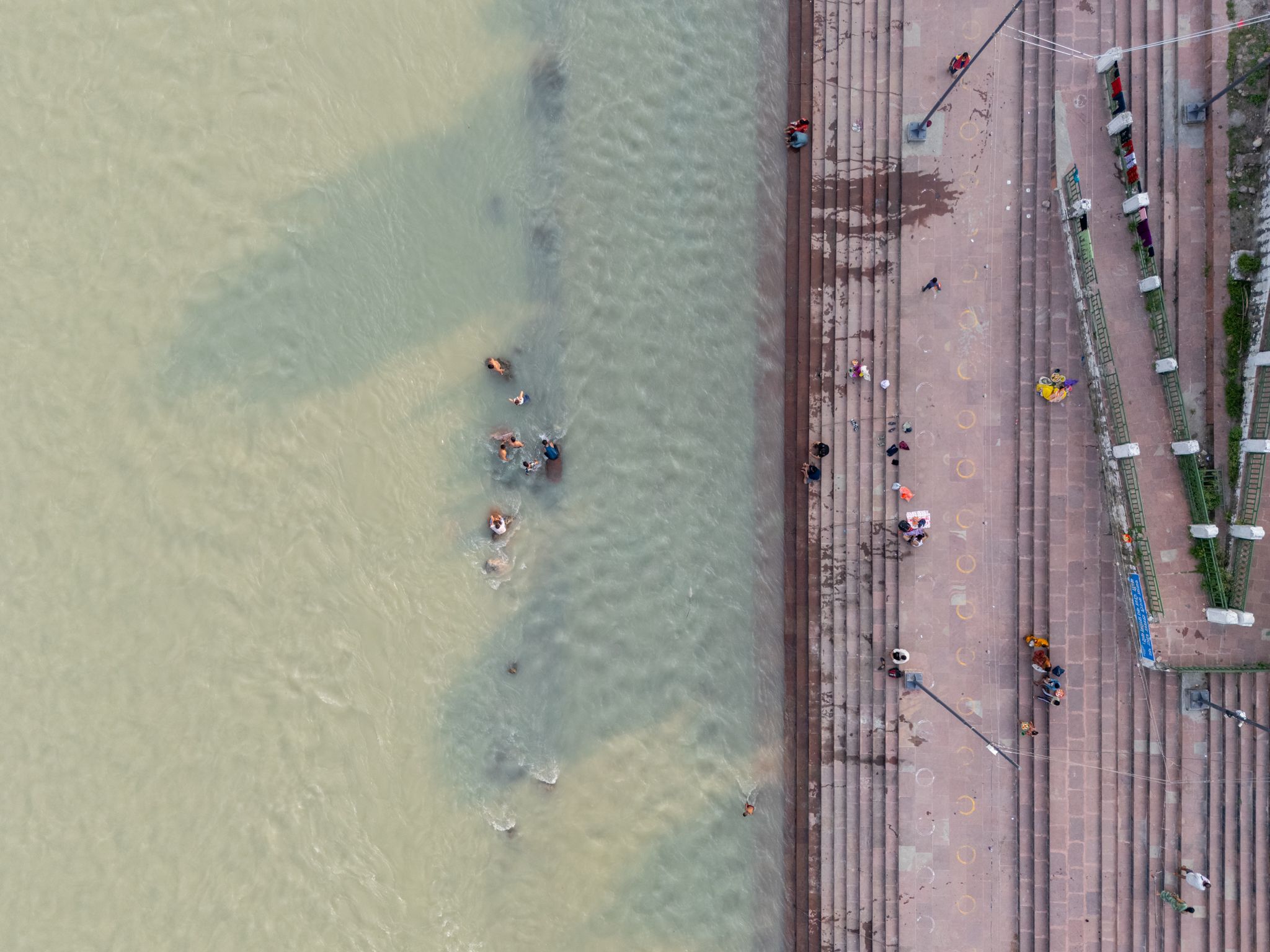 An aerial view looking straight down at the Ganga in Rishikesh, with pale green water and a few people bathing on the left, and wide stone ghat steps on the right where people walk and sit.