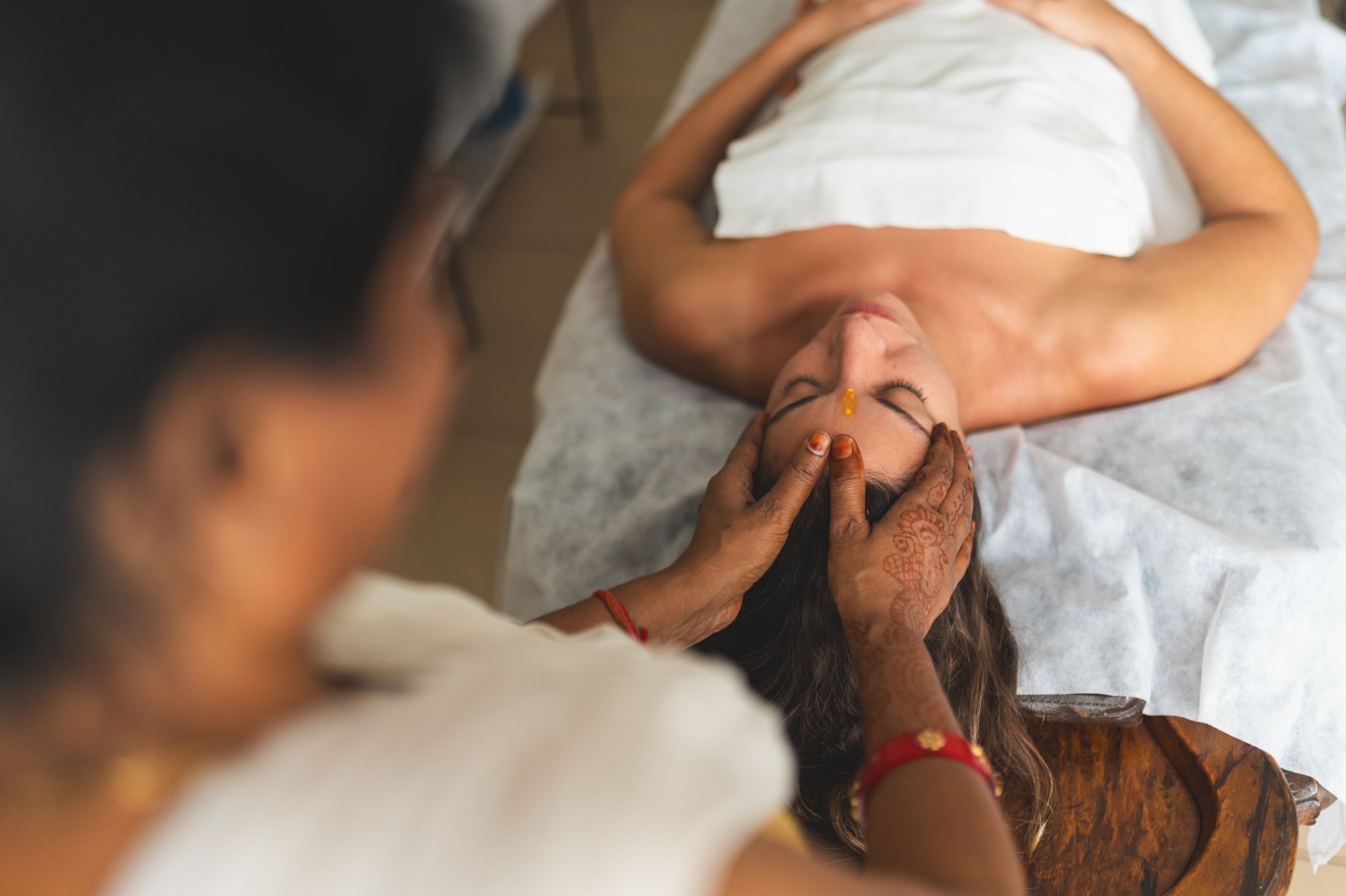 Seen from above, a woman lies on a treatment table covered with a white sheet while a therapist with hennaed hands rests her fingers on the woman's forehead and temples.
