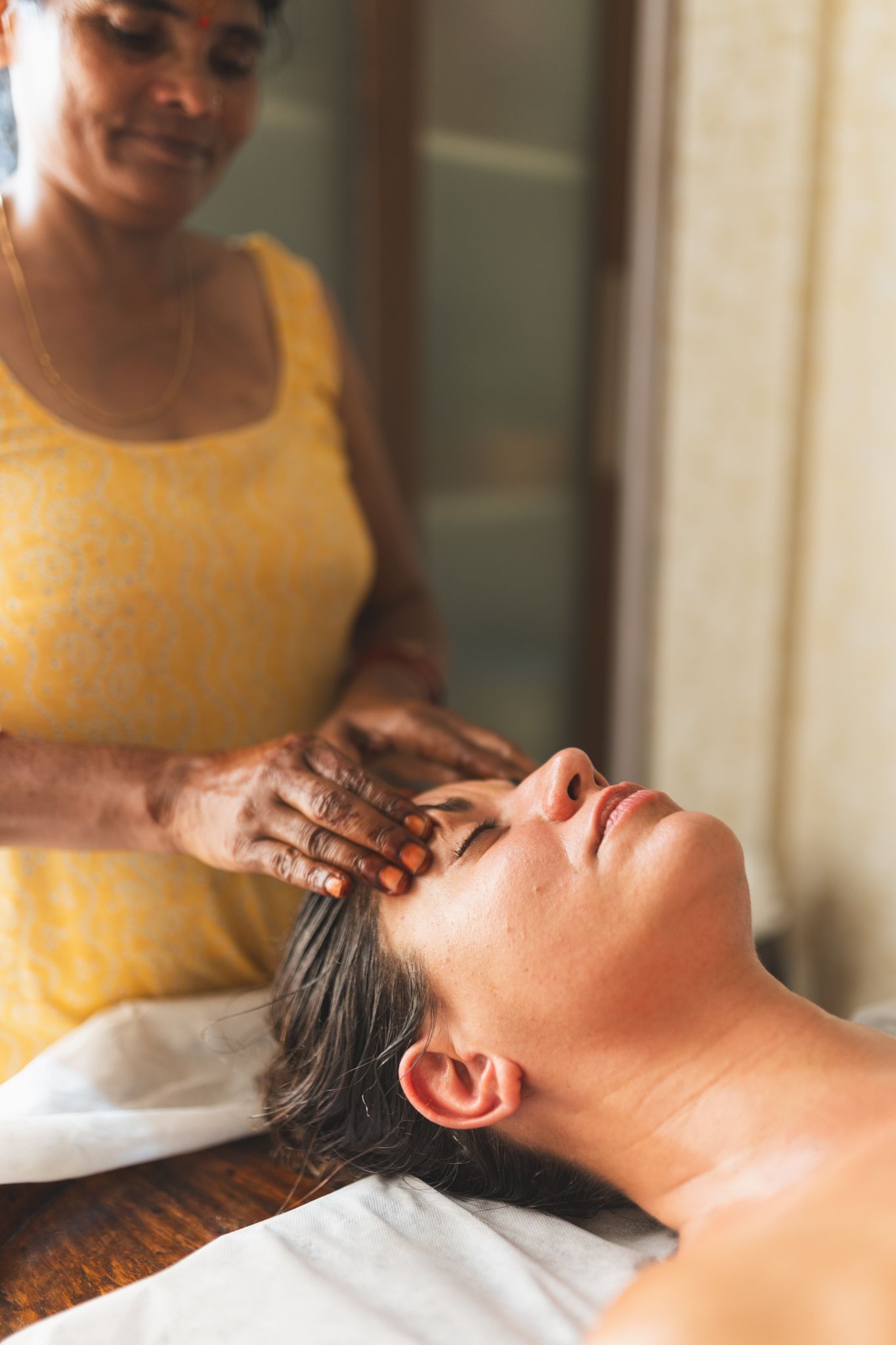 A therapist in a yellow kurta with henna on her hands massages the forehead of a woman lying with her eyes closed on a treatment table.