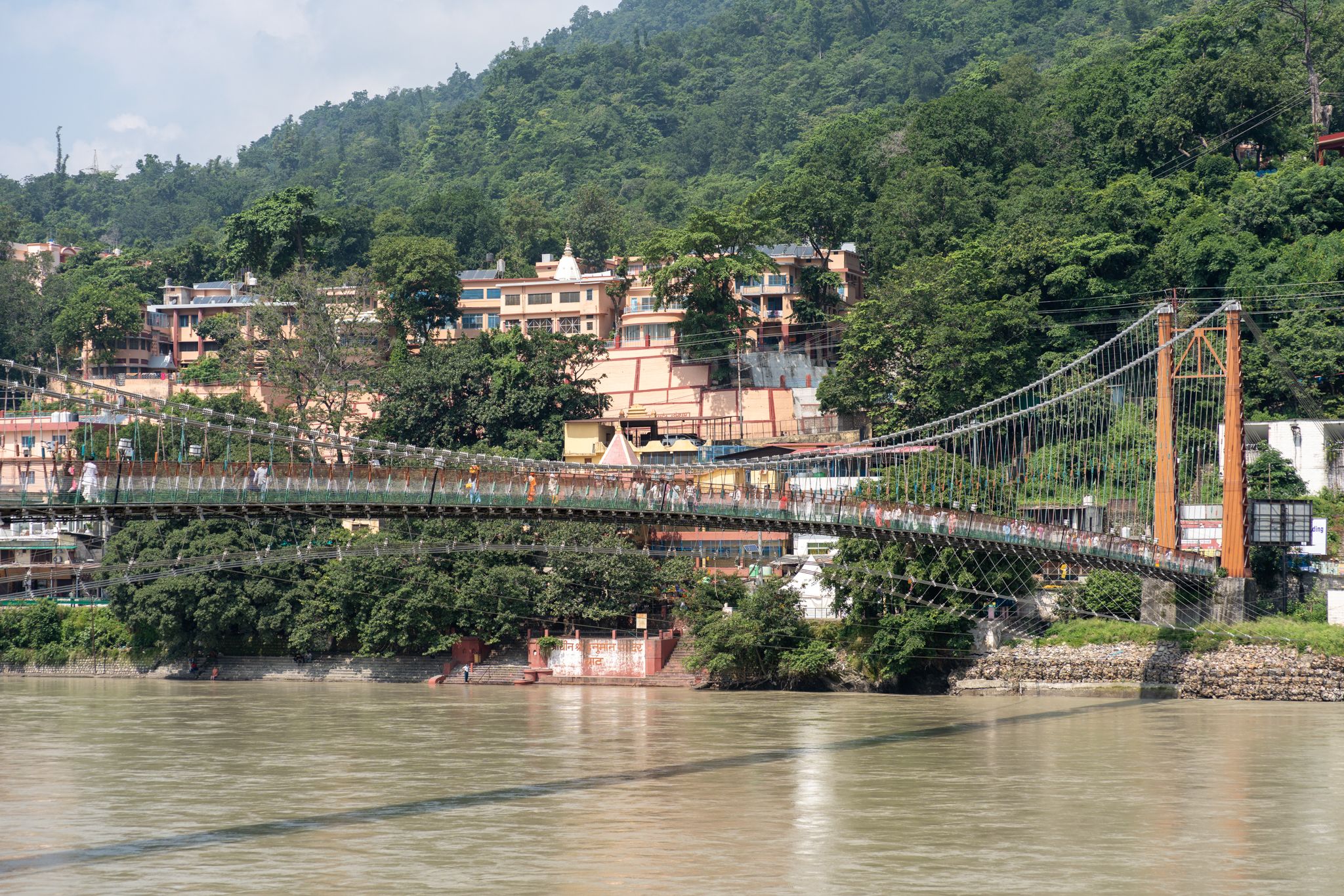 An orange-towered suspension bridge crossing the wide Ganga in Rishikesh, with temple buildings and forested hills on the far bank.